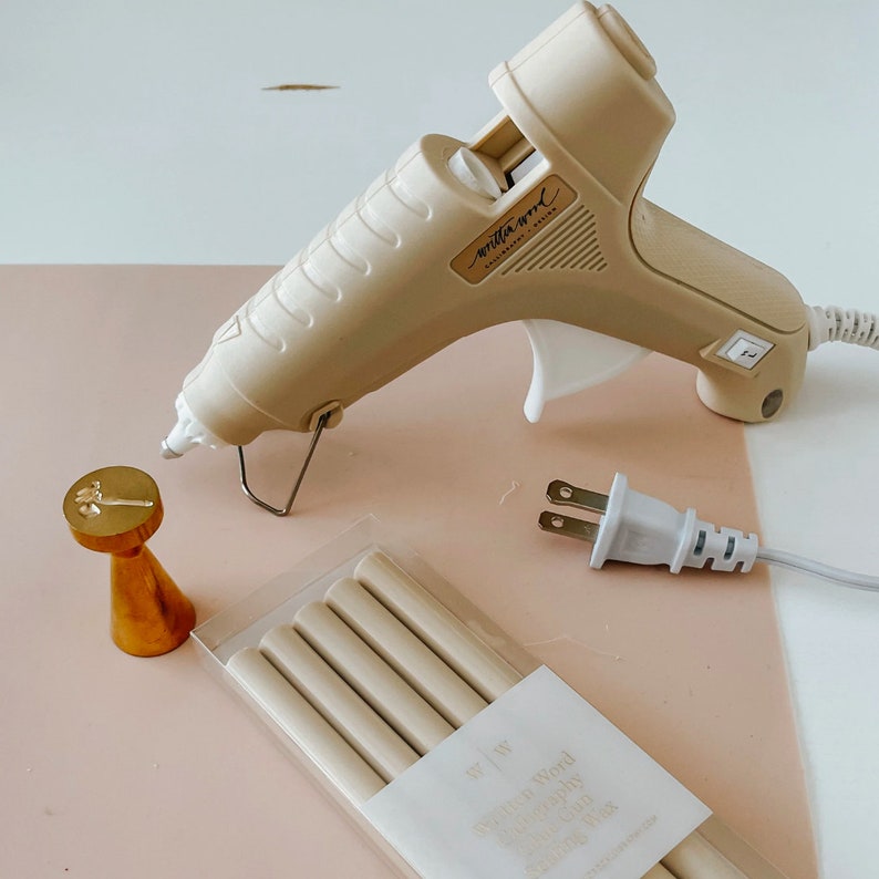 May include: A beige hot glue gun with a white plug and cord. There are six beige glue sticks in a clear plastic package with the text "Switch & Wood" and "High Quality Glue Sticks" on the package. There is also a gold metal stamp with a floral design.