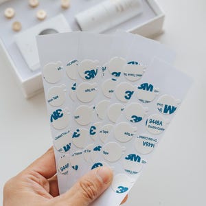 Könnte beinhalten: Eine Hand hält mehrere Blätter weißer Klebeaufkleber mit blauem 3M-Logo und dem Text "9448A Double Coated". Die Aufkleber sind rund mit einer kleinen Lasche. Im Hintergrund befindet sich eine weiße Box mit Holzscheiben.