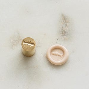 May include: A brass wax seal stamp and a salmon-colored wax seal with a fish design. The stamp is cylindrical, and the wax seal is round with a raised border. The fish is a simple, stylized outline.
