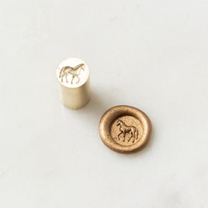 May include: Two brass horse-themed wax seal stamps. One is cylindrical, the other is a circular stamp with a raised horse design. The stamps are on a white marble surface.