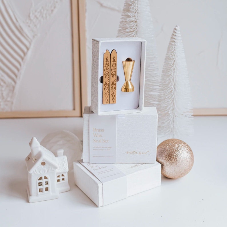 A white box with the text "Brass Wax Seal Set" on the front. The box contains two gold wax seal sticks and a brass wax seal stamp. The box is sitting on a white surface with a white ceramic house and a gold glitter ball.