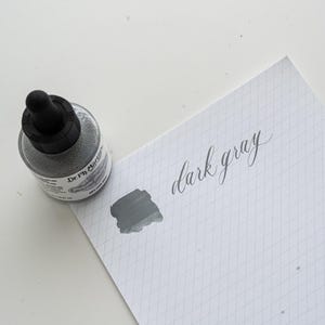 May include: A bottle of dark gray ink with a black dropper cap sits next to a piece of paper. A swatch of the ink and the words "dark gray" are written on the paper in a cursive font. The paper has a grid pattern.