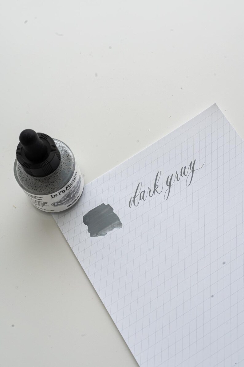 May include: A bottle of dark gray ink with a black dropper cap sits next to a piece of paper. A swatch of the ink and the words "dark gray" are written on the paper in a cursive font. The paper has a grid pattern.