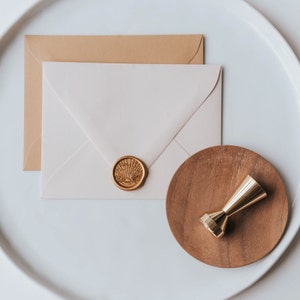 A white envelope with a gold wax seal and a brown envelope behind it. A wooden coaster with a gold metal stamp sits on a white surface.