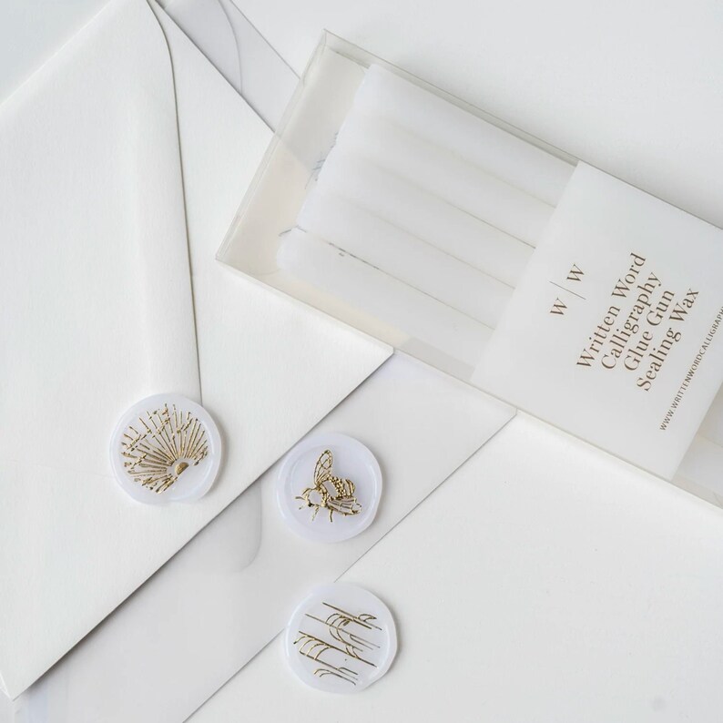 Five vellum wax sticks in a clear plastic box with the text "Written Word Calligraphy Glue Gun Sealing Wax" and the logo "W | W". Three vellum wax seals with gold designs are on a white surface.