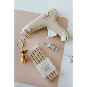 May include: A beige hot glue gun with a white power cord, two white wax seals, a gold metal stand, and a package of beige glue sticks labeled "Written Word Calligraphy Glue Gun Sealing Wax" with the website "www.writtenwordcalligraphy.com".