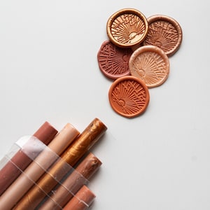 May include: Assortment of six wax seals in various shades of brown and gold, featuring a sunburst design. The seals are arranged in a semi-circle on a white surface. A bundle of six wax sticks in various shades of brown and gold are in the lower left corner of the image.