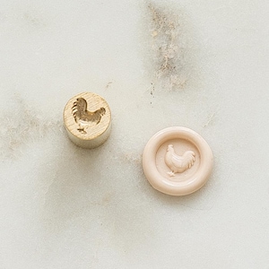 May include: A wooden stamp and a wax seal, both with a rooster design. The stamp is cylindrical and made of light-coloured wood. The wax seal is round and light pink, with the rooster in relief.