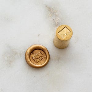 Puede incluir: Un sello de cera dorado y un mango de madera. El sello tiene un diseño circular con un icono de sobre. El sello de cera está prensado en cera dorada, creando un diseño de sobre en relieve.