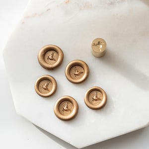 May include: Gold-colored wax seals with a bird design. The seals are round with a raised border and a detailed image of a bird in flight. A matching stamp is also present, all on a white marble surface.