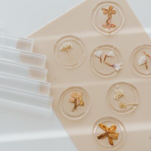 Circular clear wax seals with dried flowers encased within, displayed on a pale beige surface. The flowers are in various colors, including yellow, orange, and white. Clear sealing wax sticks are visible on the left side of the image.
