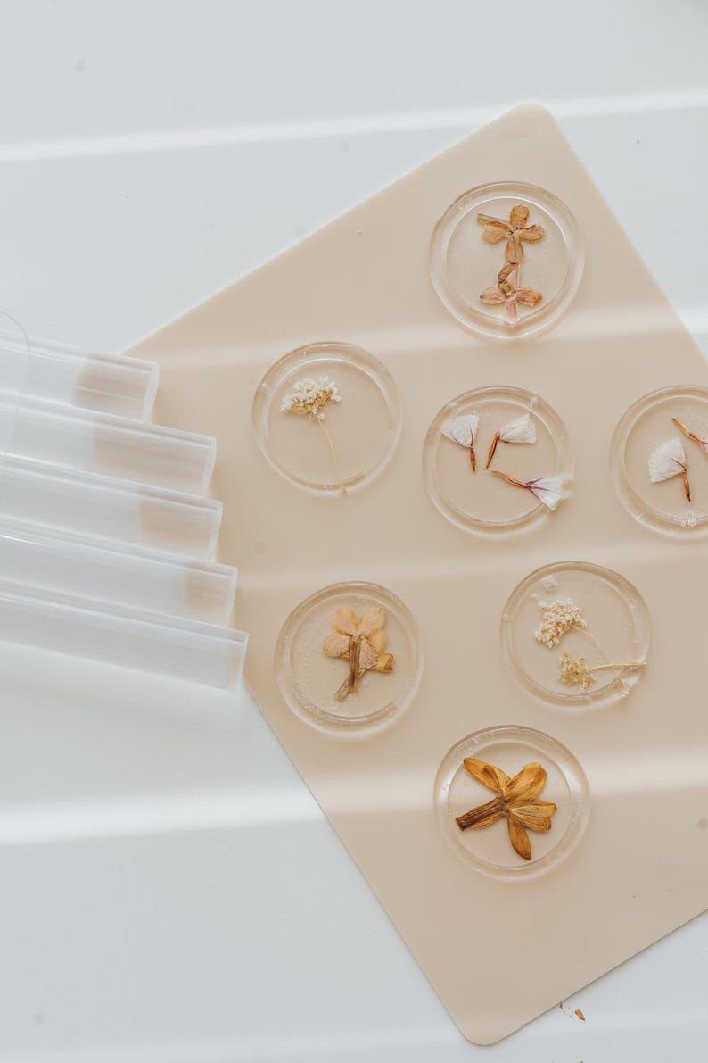 Circular clear wax seals with dried flowers encased within, displayed on a pale beige surface. The flowers are in various colors, including yellow, orange, and white. Clear sealing wax sticks are visible on the left side of the image.