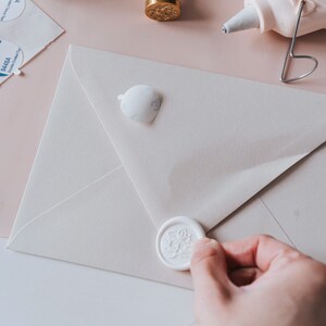 A white envelope with a white wax seal featuring a floral design. The seal is being pressed onto the envelope.