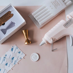 A set of wax seal supplies including a gold metal stamp, a white wax seal, a glue gun, and a pack of white sealing wax sticks. The wax sticks are labelled "Written Word Calligraphy Glue Gun Sealing Wax".