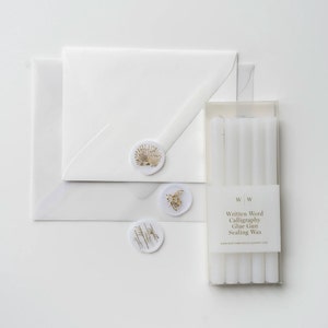 A set of white sealing wax sticks in a clear box with the text "Written Word Calligraphy Glue Gun Sealing Wax" and the website address "www.writtenwordcalligraphy.com". The box is sitting on a white surface with two white envelopes and three wax seals with gold designs.