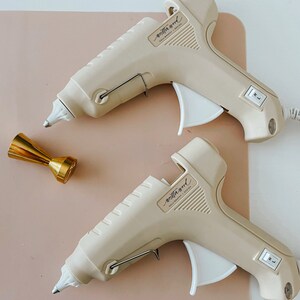 May include: Two beige hot glue guns with the brand name "mithra wood" printed on them. The glue guns are on a light pink surface.