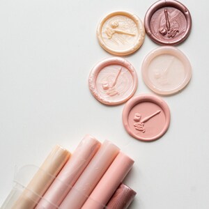 May include: An assortment of wax seals and sealing wax sticks in shades of pink, peach, and brown. The seals feature a pen and inkwell design. The wax sticks are in a clear container, ideal for crafting or stationery projects.