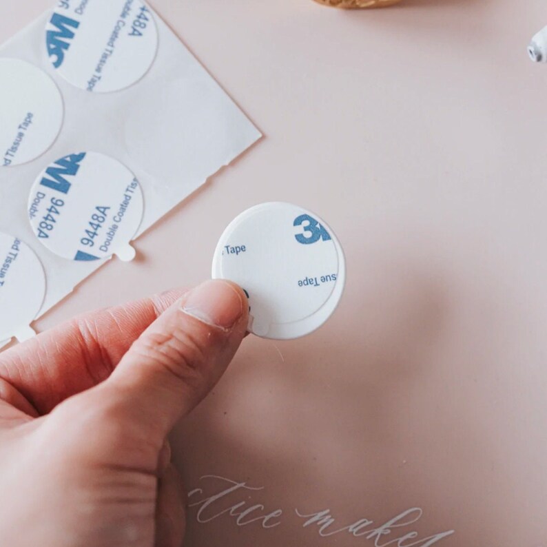 A white round adhesive circle dispenser with the text "3M" printed on it. The adhesive circle is being held by a hand.