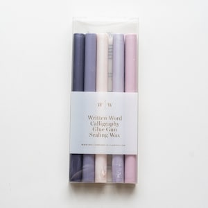 May include: A set of five sealing wax sticks in a clear plastic pack. The sticks are in shades of purple, pink, and white. The pack has a label that reads "Written Word Calligraphy Glue Gun Sealing Wax" and "www.writtenwordcalligraphy.com".