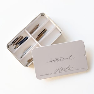 May include: A white metal box with a lid that says "written word" and "This container belongs to" with a line for a name. The lid is open and the box contains several calligraphy nibs.