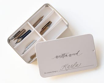 May include: A white metal box with a lid that says "written word" and "This container belongs to" with a line for a name. The lid is open and the box contains several calligraphy nibs.