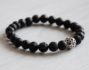 May include: A black lava rock beaded bracelet with a silver charm. The charm has a textured, circular design.
