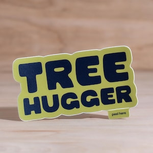 Vinyl sticker that says 'tree hugger' in all caps chunky dark navy blue font with green background and a peel here tab on a wood backdrop.