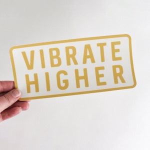 May include: White rectangular sticker with a gold border and the text "VIBRATE HIGHER" in gold letters.