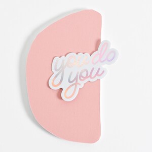 May include: A white sticker with iridescent lettering that reads "you do you" on a pink background.