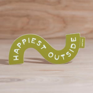 May include: A green, wavy sticker with the words "Happiest Outside" in white lettering. The sticker has a small tab that says "peel here". The sticker is on a light brown wooden surface.