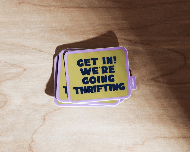 Bright green rectangular shaped sticker with rounded corners and purple border that says 'Get In! We're Going Thrifting!' and a easy peel tab on a wood background