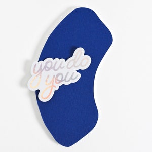 May include: A white sticker with iridescent lettering that reads "you do you" is placed on a blue curved piece of paper.