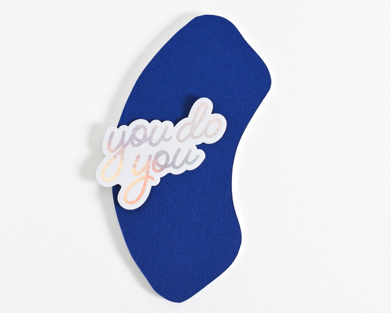 May include: A white sticker with iridescent lettering that reads "you do you" is placed on a blue curved piece of paper.
