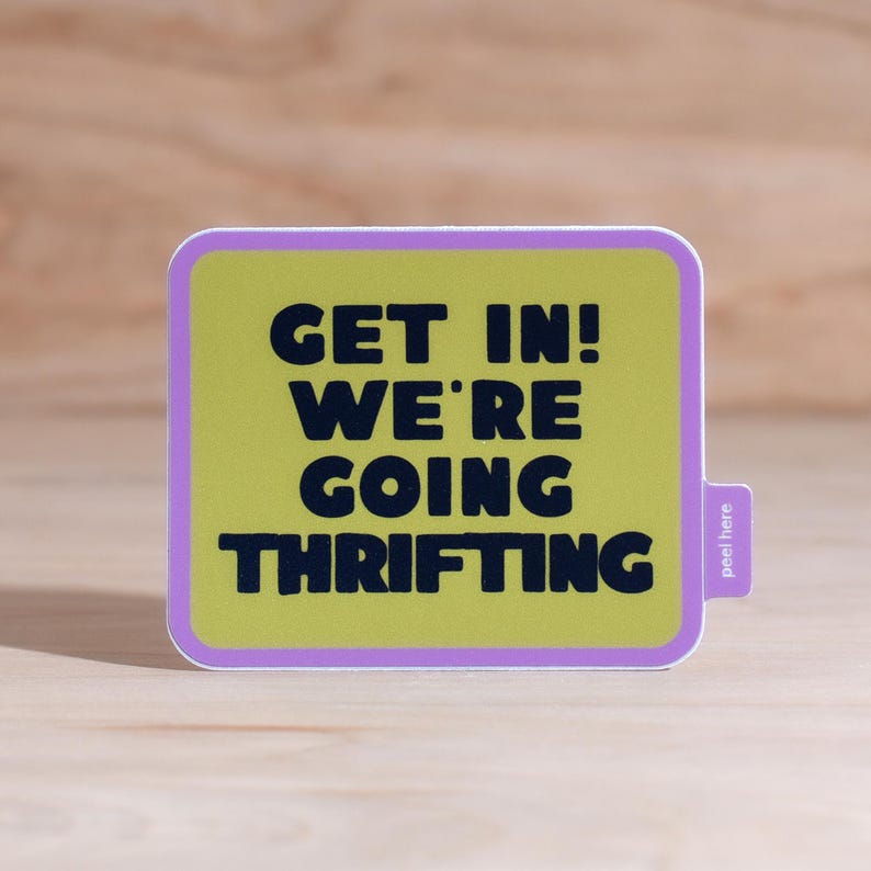 Bright green rectangular shaped sticker with rounded corners and purple border that says 'Get In! We're Going Thrifting!' in dark blue font and a easy peel tab on a wood background