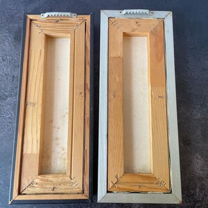 May include: Two rectangular wooden framed canvases, viewed from the back. One frame is black, the other is silver. Both have wooden supports and canvas stretched across the center. Metal sawtooth hangers are attached to the top.