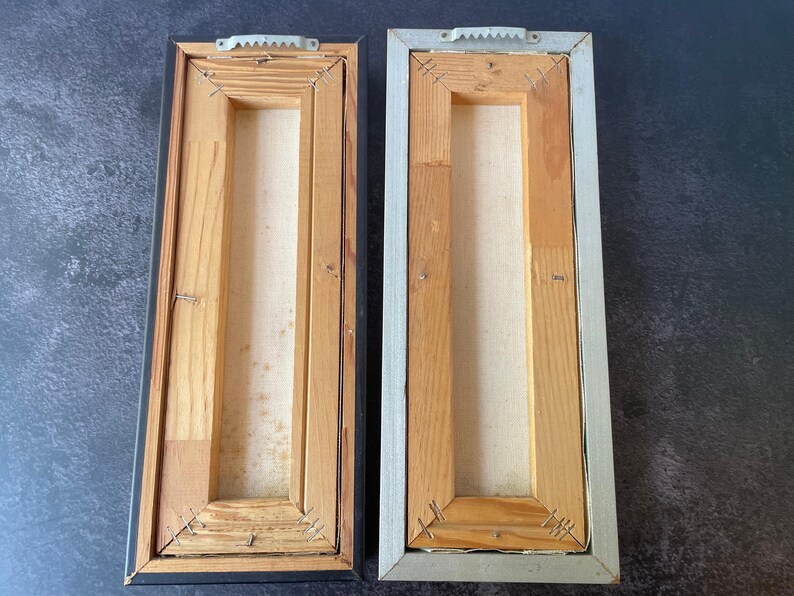 May include: Two rectangular wooden framed canvases, viewed from the back. One frame is black, the other is silver. Both have wooden supports and canvas stretched across the center. Metal sawtooth hangers are attached to the top.