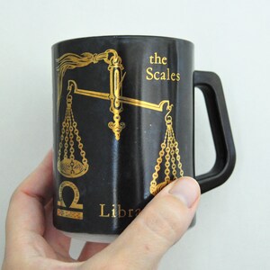 以下が含まれることがあります： 黒いセラミック製のマグカップで、金の天秤のデザインと「the Scales Libra」の文字が書かれています。