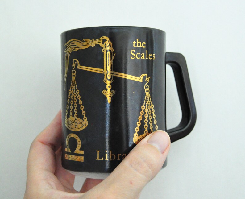 以下が含まれることがあります： 黒いセラミック製のマグカップで、金の天秤のデザインと「the Scales Libra」の文字が書かれています。