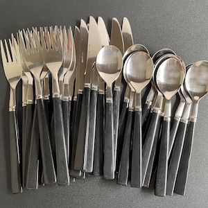 May include: A collection of stainless steel silverware with black handles. The set includes forks, knives, and spoons. The silverware is arranged on a dark surface, showcasing the polished metal and contrasting black handles.