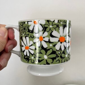 May include: A white ceramic mug with a green, black, and orange daisy pattern. The mug has a pedestal base.