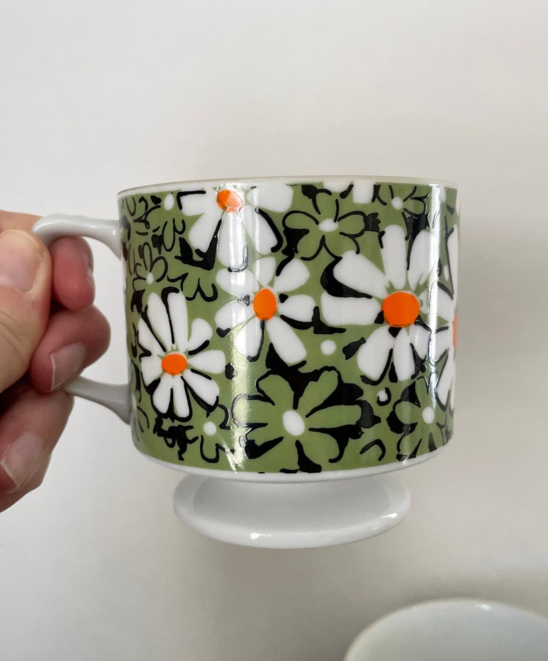 May include: A white ceramic mug with a green, black, and orange daisy pattern. The mug has a pedestal base.