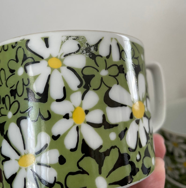 May include: A green ceramic mug with a white daisy pattern. The daisies have black outlines and yellow centers.