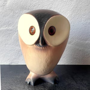 May include: A ceramic owl figurine featuring a smooth, rounded body with a gradient of brown and cream tones. The owl has large, circular cream-colored facial discs surrounding two prominent, dark orange glass eyes and a small, pointed beak.