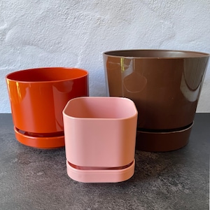 May include: Three plant pots with attached saucers. One is a large, glossy brown circular pot, one is a medium, glossy orange circular pot, and one is a smaller, matte light pink square pot with rounded corners and a distinct base design.