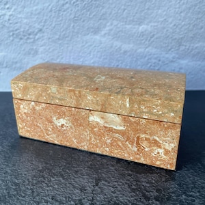 May include: A rectangular jewelry box with a hinged lid. The box is made of a reddish-brown marble with white and tan veining. The box is sitting on a dark surface.