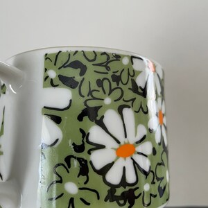 May include: A white ceramic mug with a green, black, and white floral pattern. The flowers have orange centers.