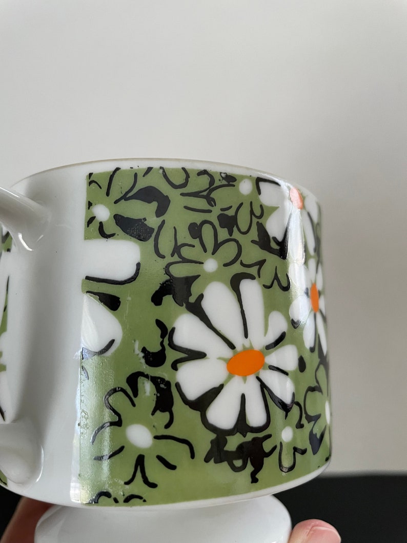 May include: A white ceramic mug with a green, black, and white floral pattern. The flowers have orange centers.