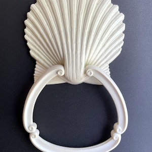 May include: White ceramic seashell towel ring. The ring is shaped like a seashell with a smooth, rounded edge. The ring is attached to a white ceramic base.