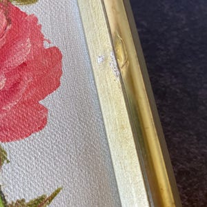 May include: Close-up of a framed floral painting. The artwork features a large red rose with green leaves against a light background. The frame is gold-colored metal and light wood. The painting is a close-up of the rose.