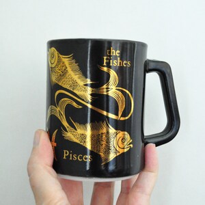 以下が含まれることがあります： 金色の魚2匹と「the Fishes Pisces」の文字が描かれた黒いセラミック製のマグカップです。